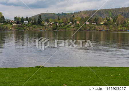 Riverside village with forested hills and cottages reflected in calm water near Nova Zivohost. Riverside village with forested hills and cottages reflected in calm water near Nova Zivohost. 135923927