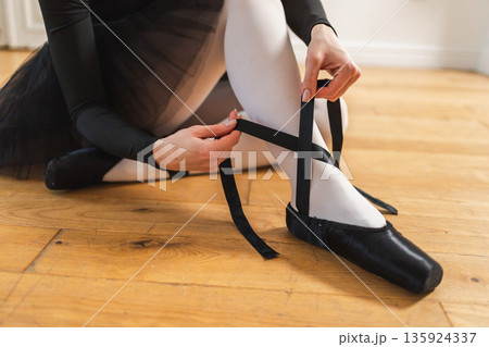 Ballerina puts on pointe shoes on leg in dance class. Classical ballet dancer woman in dance studio. Ballerina putting on tying classical pointe shoes for dance training. Ballet school concept Ballerina puts on pointe shoes on leg in dance class. Classical ballet dancer woman in dance studio. Ballerina putting on tying classical pointe shoes for dance training. Ballet school concept 135924337