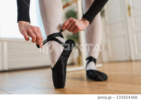 Ballerina puts on pointe shoes on leg in dance class. Classical ballet dancer woman in dance studio. Ballerina putting on tying classical pointe shoes for dance training. Ballet school concept Ballerina puts on pointe shoes on leg in dance class. Classical ballet dancer woman in dance studio. Ballerina putting on tying classical pointe shoes for dance training. Ballet school concept 135924366