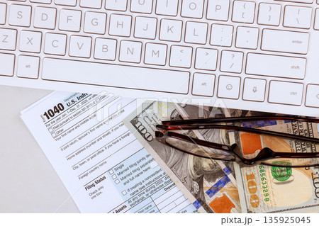 Accouter fills out tax 1040 forms with USA cash, glasses on desk while working computer during tax season Accouter fills out tax 1040 forms with USA cash, glasses on desk while working computer during tax season 135925045