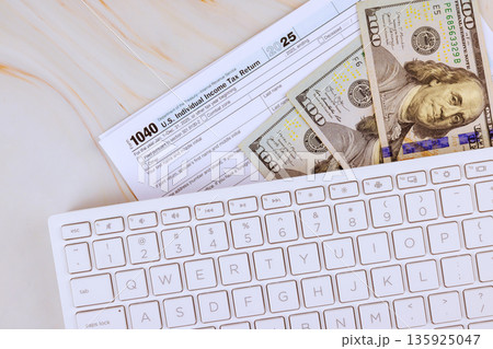 Tax 1040 return form on desk next to several one hundred dollar bills, bookkeeping work during financial tax preparations. Tax 1040 return form on desk next to several one hundred dollar bills, bookkeeping work during financial tax preparations. 135925047