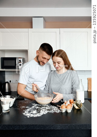 Couple prepares dessert together in a modern kitchen during the afternoon while smiling and having fun with ingredients on the counter 135925071