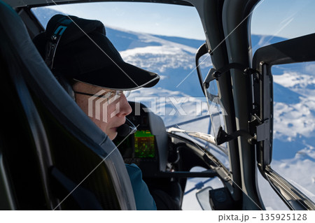 Pilot in a helicopter cockpit over snowy mountains, capturing fresh powder scenery Pilot in a helicopter cockpit over snowy mountains, capturing fresh powder scenery 135925128
