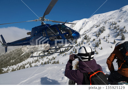 Blue helicopter in alpine snow as skiers photograph powder day with female heliskiing pilot. Blue helicopter in alpine snow as skiers photograph powder day with female heliskiing pilot. 135925129
