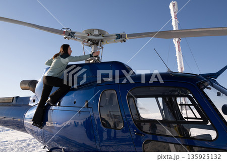 Woman climbs onto a blue helicopter in bright winter sun, preparing for a snow flight adventure Woman climbs onto a blue helicopter in bright winter sun, preparing for a snow flight adventure 135925132