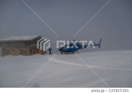 Blue helicopter on fresh powder beside a mountain lodge, ready for heliskiing adventure 135925163