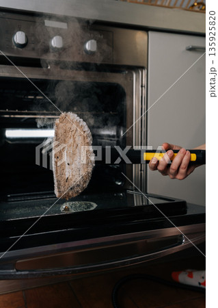 Housewife using steam cleaner with cleaning pad, generating steam and cleaning dirty stains inside kitchen oven, highlighting effective household chores and natural hygiene. Housewife using steam cleaner with cleaning pad, generating steam and cleaning dirty stains inside kitchen oven, highlighting effective household chores and natural hygiene. 135925820