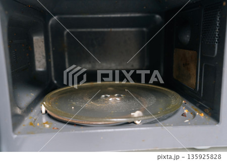 Microwave oven interior with soiled glass turntable and food residue, representing poor kitchen cleanliness, unhygienic conditions, and need for regular appliance maintenance. Microwave oven interior with soiled glass turntable and food residue, representing poor kitchen cleanliness, unhygienic conditions, and need for regular appliance maintenance. 135925828