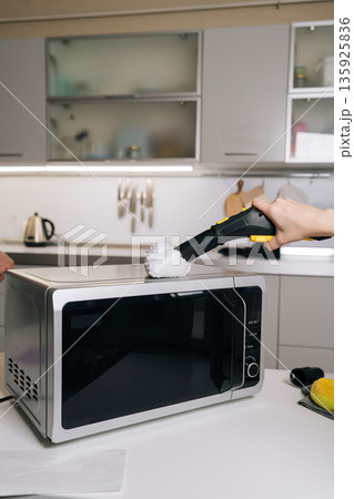 Closeup of unrecognizable housekeeper cleaning microwave top with steam cleaner, removing grease and germs for spotless kitchen appliance. Concept of domestic hygiene and routine maintenance. Closeup of unrecognizable housekeeper cleaning microwave top with steam cleaner, removing grease and germs for spotless kitchen appliance. Concept of domestic hygiene and routine maintenance. 135925836