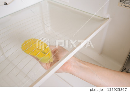 Close-up of hand cleaning refrigerator interior, scrubbing glass shelf with sponge to remove dirt and food residue, demonstrating routine domestic hygiene and upkeep. Concept of household cleanliness. Close-up of hand cleaning refrigerator interior, scrubbing glass shelf with sponge to remove dirt and food residue, demonstrating routine domestic hygiene and upkeep. Concept of household cleanliness. 135925867
