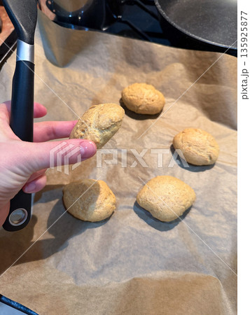 Fresh homemade cookies rest on baking paper while hand holds one cookie above the tray, warm tones and soft kitchen light highlight the golden texture 135925877