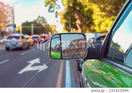 Car side mirror reflecting city traffic on urban street. Car side mirror reflects buildings and traffic on a city street during daylight. Transportation, road safety, navigation and commuting concept Car side mirror reflecting city traffic on urban street. Car side mirror reflects buildings and traffic on a city street during daylight. Transportation, road safety, navigation and commuting concept 135925894