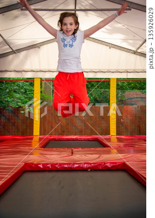 Happy girl having fun while jumping on trampoline. Sports girl jumps on a trampoline. Outdoor shot of girl jumping on trampoline, enjoys jumping in home. happy summer vacation. High quality photo Happy girl having fun while jumping on trampoline. Sports girl jumps on a trampoline. Outdoor shot of girl jumping on trampoline, enjoys jumping in home. happy summer vacation. High quality photo 135926029