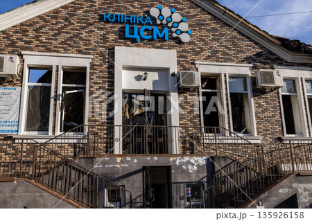 Brick clinic with Cyrillic signage shows broken windows and exterior damage; air conditioners and plants remain intact under clear blue sky. 135926158