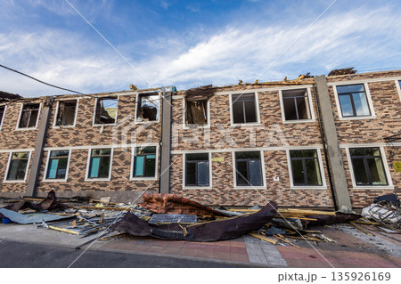 Two story brick building with patterned facade shows partial roof collapse, broken windows, and scattered debris damage suggests recent destructive event. Two story brick building with patterned facade shows partial roof collapse, broken windows, and scattered debris damage suggests recent destructive event. 135926169