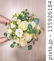 White floral bouquet held by hands, featuring roses, lisianthus, and greenery against a light wooden background 135926184
