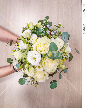 White floral bouquet held by hands, featuring roses, lisianthus, and greenery against a light wooden background 135926184