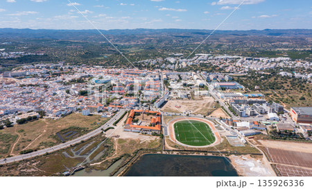Tavira shows its layout with buildings, fields, and a sports field. The view captures the blend of nature and urban areas in the Algarve region during daytime Tavira shows its layout with buildings, fields, and a sports field. The view captures the blend of nature and urban areas in the Algarve region during daytime 135926336