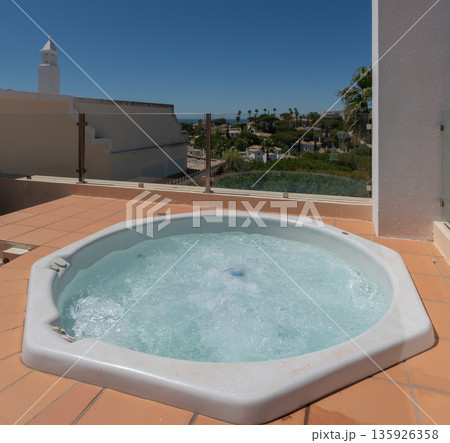 A hot tub sits on a rooftop terrace, surrounded by clear sky and palm trees, offering a view of the ocean and nearby landscape in bright weather 135926358