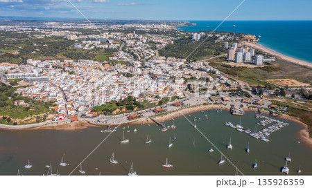 Aerial view of coastal town with vibrant buildings, boats in harbor, and scenic landscape showcasing tourism in a picturesque destination. Alvor Portugal Algarve Aerial view of coastal town with vibrant buildings, boats in harbor, and scenic landscape showcasing tourism in a picturesque destination. Alvor Portugal Algarve 135926359