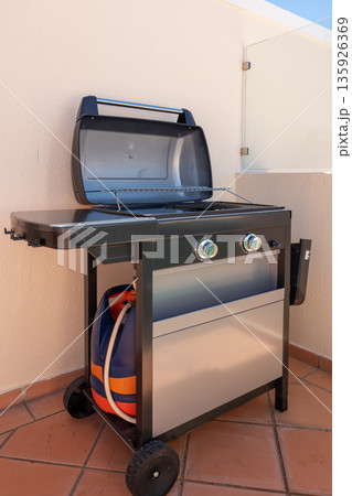 A grill sits on a patio terrace with its lid open, revealing the cooking surface and gas connections. It is positioned for outdoor cooking during the day A grill sits on a patio terrace with its lid open, revealing the cooking surface and gas connections. It is positioned for outdoor cooking during the day 135926369
