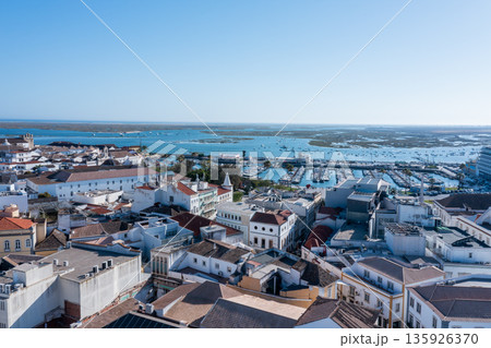 Tourists explore Faro's Old Town while boats float in the blue waters of Ria Formosa. The clear sky highlights the beauty of the Algarve region 135926370