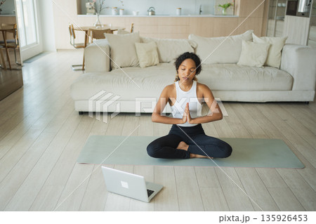 Yoga mindfulness meditation online. African woman practicing yoga with online lessons in laptop at home. Woman sitting in lotus pose meditating relaxing. Girl doing breathing practice. Yoga at home Yoga mindfulness meditation online. African woman practicing yoga with online lessons in laptop at home. Woman sitting in lotus pose meditating relaxing. Girl doing breathing practice. Yoga at home 135926453
