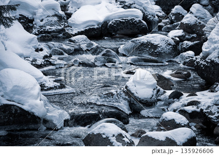 A tranquil winter scene features a cold river flowing through rocks covered in snow. The icy landscape captures the serene beauty of winter in the Czech Republic. A tranquil winter scene features a cold river flowing through rocks covered in snow. The icy landscape captures the serene beauty of winter in the Czech Republic. 135926606