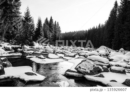 The Jizera River flows peacefully on a winter evening, surrounded by snow-covered rocks and tall evergreen trees. The serene landscape captures the beauty of the border area in winter. The Jizera River flows peacefully on a winter evening, surrounded by snow-covered rocks and tall evergreen trees. The serene landscape captures the beauty of the border area in winter. 135926607