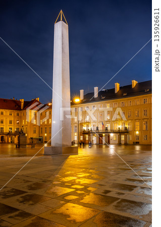 The Mrakotin Monolith stands tall at Prague Castle, surrounded by historical buildings, as warm light casts dramatic shadows on the cobblestone ground under the night sky. The Mrakotin Monolith stands tall at Prague Castle, surrounded by historical buildings, as warm light casts dramatic shadows on the cobblestone ground under the night sky. 135926611