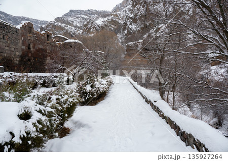 Snow-covered path leads toward stone walls of Geghard Monastery, framed by bare trees and steep rocky cliffs in muted winter light Snow-covered path leads toward stone walls of Geghard Monastery, framed by bare trees and steep rocky cliffs in muted winter light 135927264