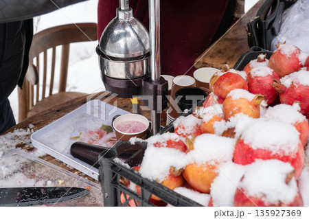 Fresh pomegranates covered with snow sit beside manual juice press, paper cups at outdoor stand, vivid red fruit contrasts with metal tools 135927369