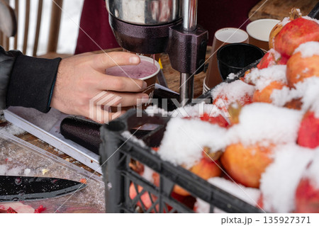 Fresh pomegranate juice is poured into a paper cup at street stand, bright red fruit covered with snow contrasts with metal press and winter textures 135927371