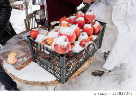 Fresh pomegranates covered with snow sit beside manual juice press, paper cups at outdoor stand, vivid red fruit contrasts with metal tools 135927373