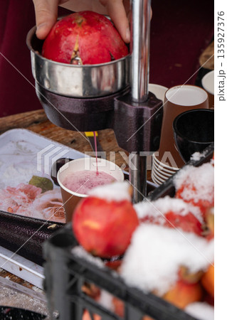 Fresh pomegranate juice flows from manual press into paper cup, rich red color contrasts with metal equipment, snow-covered fruit Fresh pomegranate juice flows from manual press into paper cup, rich red color contrasts with metal equipment, snow-covered fruit 135927376