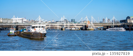 Boats float on the Thames River near Hungerford Bridge in London. People walk across the bridge enjoying the day. The city skyline appears in the background under a blue sky. Boats float on the Thames River near Hungerford Bridge in London. People walk across the bridge enjoying the day. The city skyline appears in the background under a blue sky. 135927819