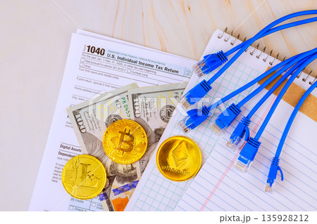 Tax return form lies with dollar cash cryptocurrency coins along with blue cables in accounting office setting. Tax return form lies with dollar cash cryptocurrency coins along with blue cables in accounting office setting. 135928212