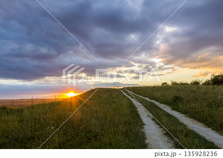 Beautiful landscape at sunset or sunrise, narrow ground road stretching through green grassy blooming meadow to distant horizon lit by bright rays of yellow low setting sun under cloudy evening sky. Beautiful landscape at sunset or sunrise, narrow ground road stretching through green grassy blooming meadow to distant horizon lit by bright rays of yellow low setting sun under cloudy evening sky. 135928236