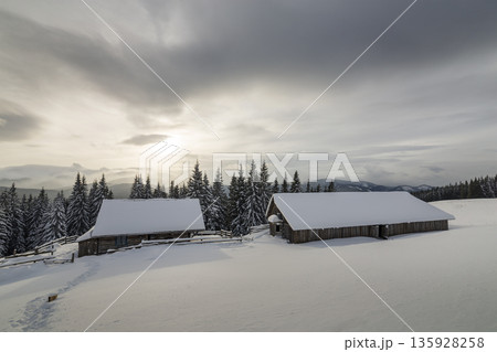 Winter mountain landscape. Old wooden houses on snowy clearing on background of mountain ridge, spruce forest and cloudy sky. Happy New Year and Merry Christmas card. Winter mountain landscape. Old wooden houses on snowy clearing on background of mountain ridge, spruce forest and cloudy sky. Happy New Year and Merry Christmas card. 135928258