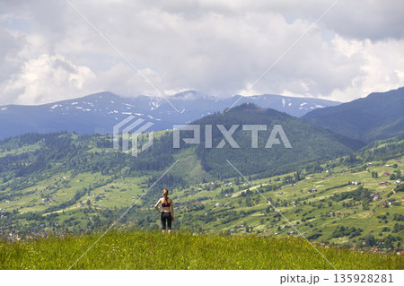 Back view of slim young woman standing on grassy valley on background of green mountains on sunny summer day. Back view of slim young woman standing on grassy valley on background of green mountains on sunny summer day. 135928281