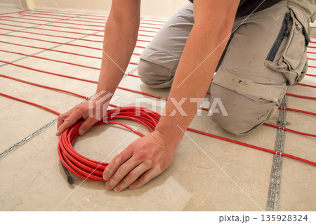 Electrician installing heating red electrical cable wire on cement floor in unfinished room. Renovation and construction, comfortable warm home concept. 135928324