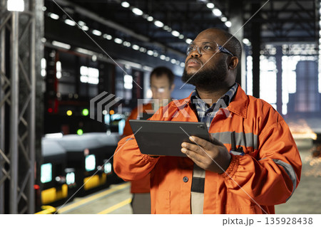 Black engineering supervisor in protective gear overseeing welding activity with tablet, reflecting heavy labor and the automation development of the industrial factory as sparks rise. Black engineering supervisor in protective gear overseeing welding activity with tablet, reflecting heavy labor and the automation development of the industrial factory as sparks rise. 135928438