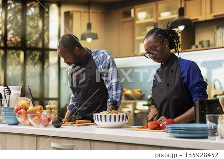 Black young couple chopping fresh bell peppers and carrots for tasty salad, preparing food at home. Man and woman preparing bio vegetables for a vegetarian lunch, joyful activity. 135928452