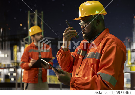 Offshore rig expert uses walkie talkie to coordinate safety protocols with logistics staff members at night. African american drilling barge radio operator talks into handheld device, supervising deck Offshore rig expert uses walkie talkie to coordinate safety protocols with logistics staff members at night. African american drilling barge radio operator talks into handheld device, supervising deck 135928463
