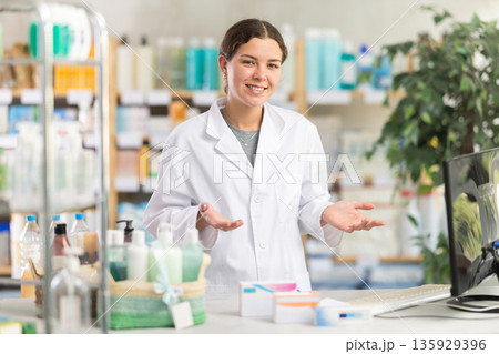Skilled young woman pharmacist standing at prescription counter and inviting clients at pharmacy shop 135929396