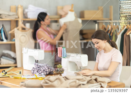 Woman tailor sews clothes in sewing machine. Companion in background Woman tailor sews clothes in sewing machine. Companion in background 135929397