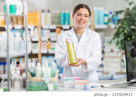 Skilled happy young woman pharmacist standing at prescription counter and offering shampoo or shower gel to clients at pharmacy Skilled happy young woman pharmacist standing at prescription counter and offering shampoo or shower gel to clients at pharmacy 135929413