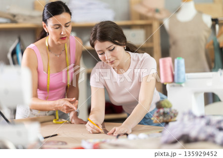 Manager of sewing workshop teaches a young girl seamstress to draw using patterns Manager of sewing workshop teaches a young girl seamstress to draw using patterns 135929452