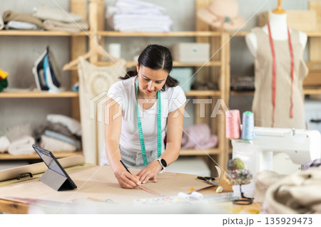 Woman tailor draws dress pattern of product on paper, tablet on table. 135929473