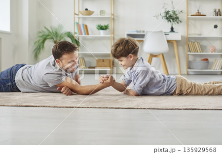 Happy dad and little son are lying on the floor and having an arm wrestling contest Happy dad and little son are lying on the floor and having an arm wrestling contest 135929568